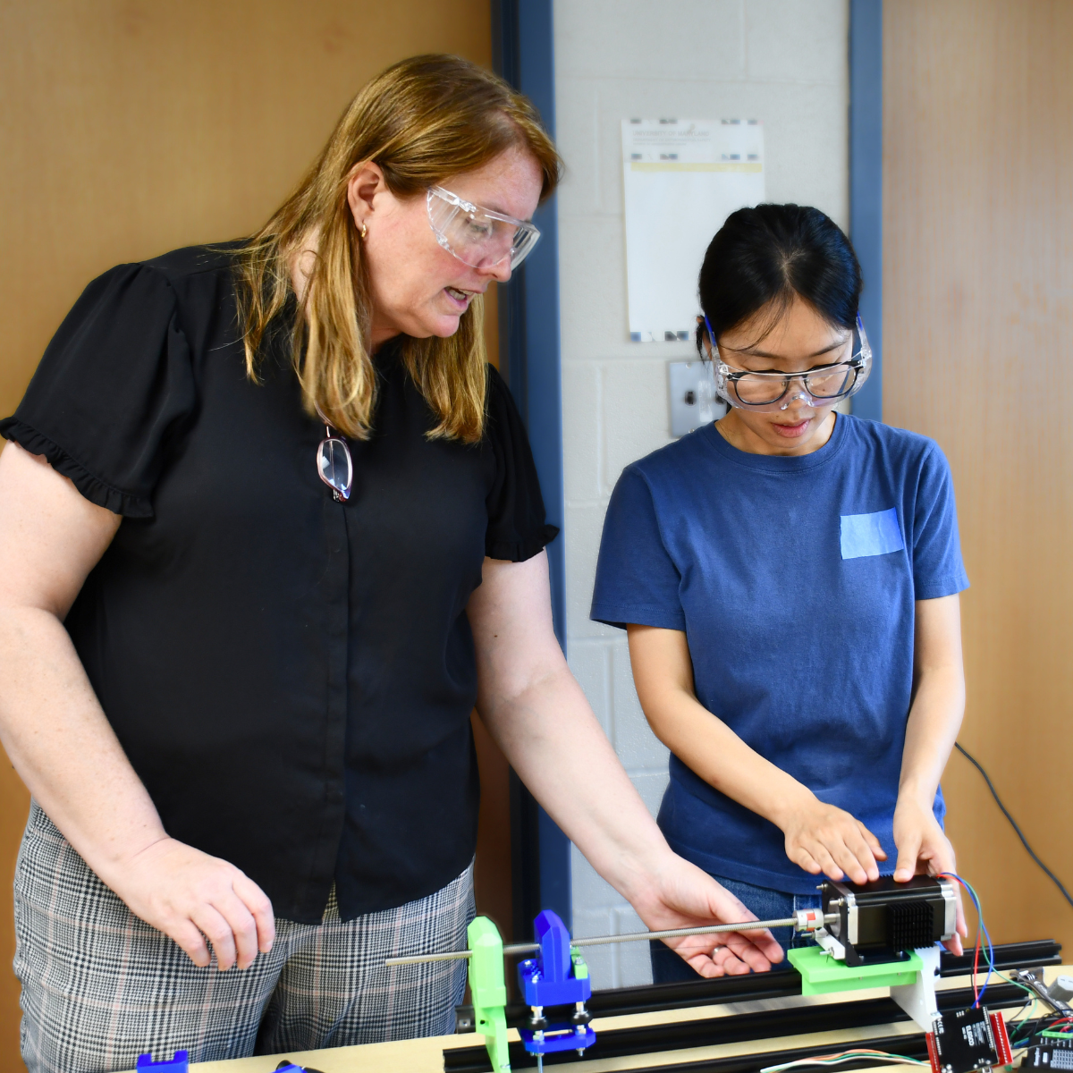 A graduate student helps guide an RET teacher through an elastocaloric cooling experiment. The two are standing together and are examining the prototype, which is sitting on a table in front of them.