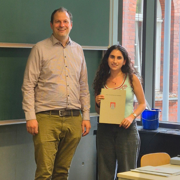 Yara Daher is standing, holding her program certificate in a classroom at Hamburg University of Technology. Standing next to her is Professor Arne Speerforck.